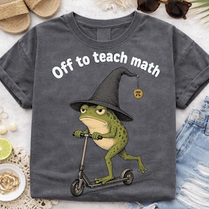 May include: Dark gray t-shirt with the text "Off to teach math." A cartoon frog wearing a witch hat and riding a scooter is featured on the front. A pi symbol hangs from the hat.