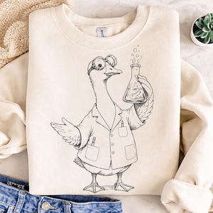 May include: A cream-colored crewneck sweatshirt featuring a black line art illustration of a goose dressed as a scientist. The goose wears goggles and a lab coat with pens in the pockets, holding a bubbling Erlenmeyer flask with a small fish inside.