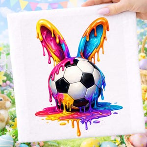 May include: A white square towel featuring a soccer ball with black and white pentagons, topped with colorful bunny ears. The ears and the ball are dripping with vibrant rainbow-colored paint, creating a unique and playful design.