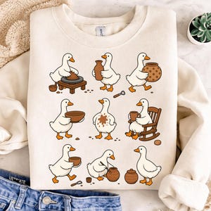 May include: Cream-colored sweatshirt featuring a whimsical design of cartoon geese engaged in pottery-making activities. The geese are depicted working with clay, holding mugs, and relaxing in a rocking chair. The design is playful and artistic.