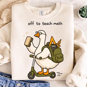 May include: A cream-colored crewneck sweatshirt featuring a graphic of a white goose wearing glasses, riding a green scooter, and carrying a green backpack filled with a yellow triangle ruler, a compass, and a calculator, with the text off to teach math.