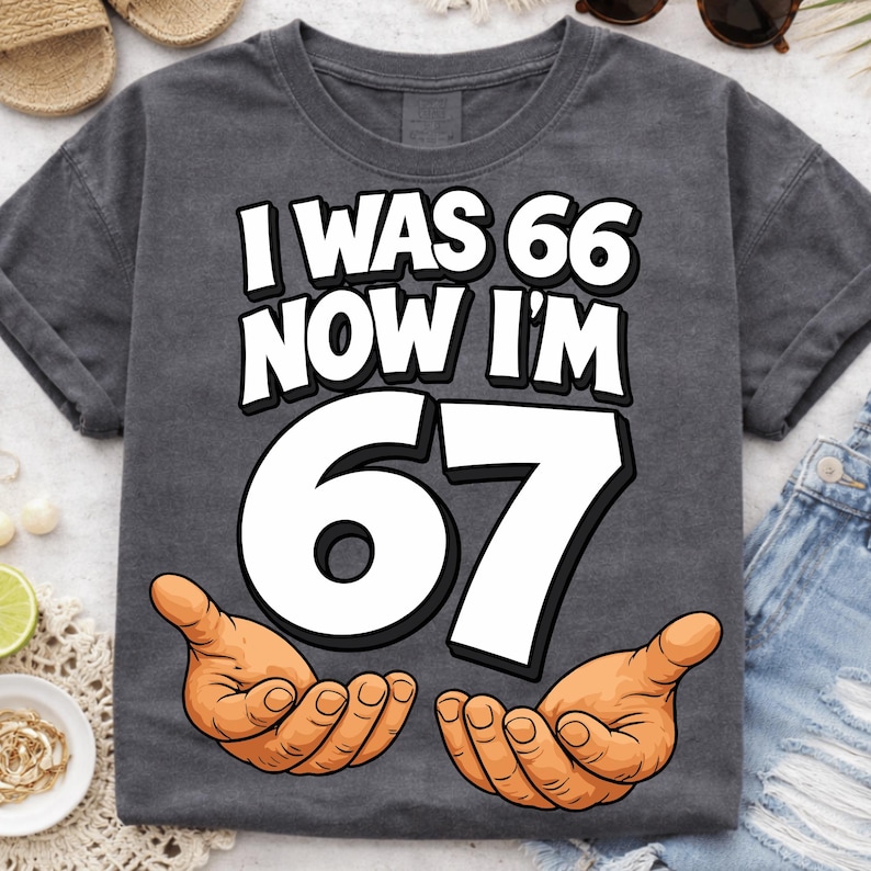 May include: Dark gray t-shirt with the text "I WAS 66 NOW I'M 67" in white, with a large white "67" and two cartoon hands. The shirt is a casual style.