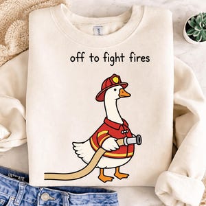 Könnte beinhalten: Cremefarbenes Sweatshirt mit dem Text "off to fight fires". Eine Cartoon-Gans in roter Feuerwehruniform und Hut hält einen Schlauch. Die Gans ist weiß mit einem orangefarbenen Schnabel und Füßen.