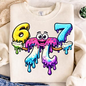 May include: Cream-colored sweatshirt featuring a colorful cartoon design of the number 67. The numbers are designed to look like melting donuts with yellow, blue, pink, and purple frosting. The design has a smiling face.