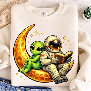 May include: Cream-colored sweatshirt featuring a cartoon illustration of an astronaut and a green alien sitting on a crescent moon. The astronaut is reading a book. The moon is yellow and orange with stars.