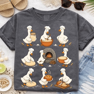 Könnte beinhalten: Dunkelgraues T-Shirt mit einem lustigen Design von Gänsen, die Brot backen. Die Gänse tragen Kochmützen und werden in verschiedenen Phasen der Brotherstellung gezeigt, vom Mischen der Zutaten bis zum Backen in einem Ziegelofen. Das Design umfasst Weizenähren und Brot.