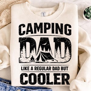 May include: Cream-colored sweatshirt with the text "CAMPING DAD" in a distressed black font. The word "DAD" features a camping scene with trees, a tent, and a campfire. The text "LIKE A REGULAR DAD BUT COOLER" is below.