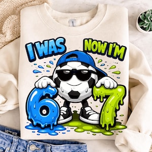 May include: Cream-colored sweatshirt with a soccer ball graphic wearing sunglasses and a blue cap. The graphic includes the text "I WAS" and "NOW I'M" with the numbers 6 and 7 in blue and green.