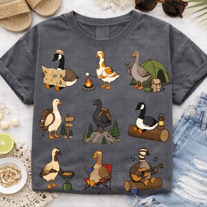 Puede incluir: Camiseta gris oscuro con dibujos animados de gansos acampando. Los gansos se muestran con mochilas, mapas, fogatas e instrumentos musicales. La camiseta tiene un corte holgado y mangas enrolladas.