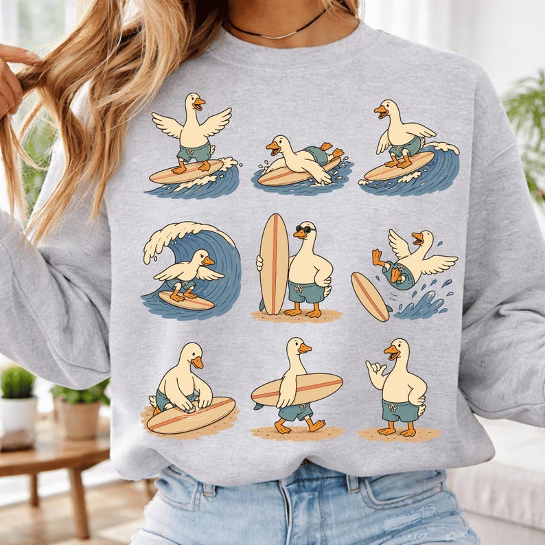 Pode incluir: Uma camisola cinzenta clara de gola redonda com uma impress&atilde;o gr&aacute;fica de nove patos de desenhos animados em cal&ccedil;&otilde;es de banho azuis a surfar ondas ou a transportar pranchas de surf. Os patos s&atilde;o retratados em v&aacute;rias poses, incluindo ondas.