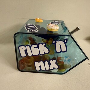 Könnte beinhalten: Ein klarer, blauer, angewinkelter Süßigkeitenbehälter mit den Worten "Pick 'n' Mix" in weißen Buchstaben. Der Behälter enthält verschiedene Süßigkeiten, darunter Gummienten und -ringe. Kleine dekorative Donuts sind oben.