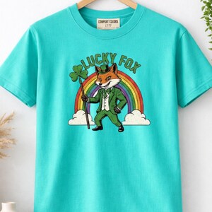 May include: A turquoise Comfort Colors t-shirt featuring a graphic of a fox in a green suit and hat, holding a shamrock and staff in front of a rainbow. The text "LUCKY FOX" is arched above the fox.