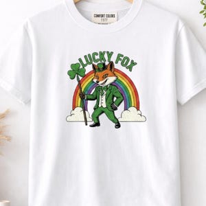 May include: White t-shirt with a graphic of a fox dressed in a green suit and hat, holding a shamrock. The text "LUCKY FOX" is above the fox, with a rainbow in the background. The shirt is a Comfort Colors 1177.