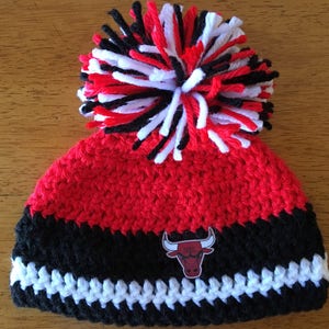 Pode incluir: Chap&eacute;u de croch&ecirc; vermelho e preto com um pompom branco e um logotipo de touro preto e vermelho.