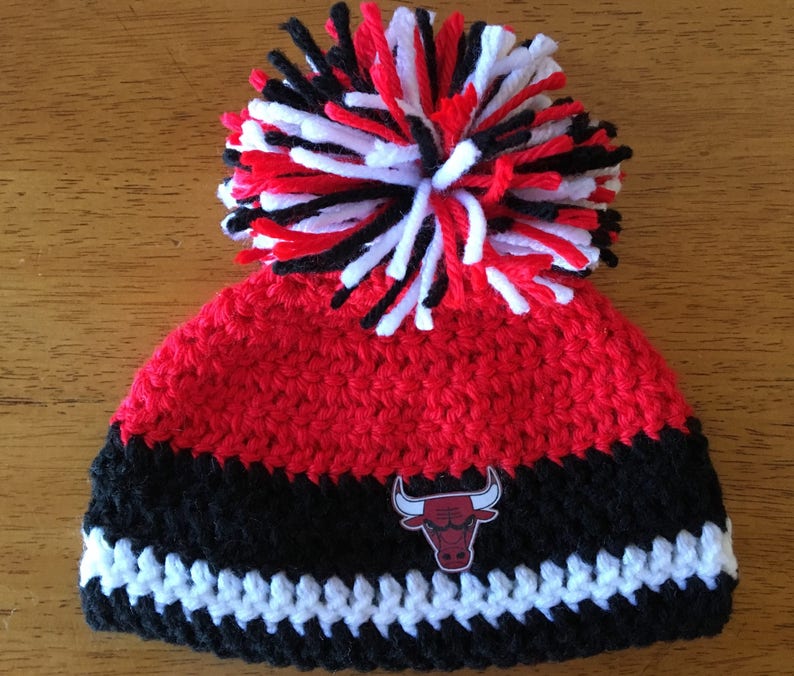 Pode incluir: Chap&eacute;u de croch&ecirc; vermelho e preto com um pompom branco e um logotipo de touro preto e vermelho.