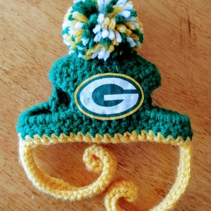May include: A handmade, green and yellow crocheted hat with a pom-pom and the Green Bay Packers logo. The hat features a green body, yellow trim, and a white and yellow pom-pom. The logo is an oval with a white background and a green "G".