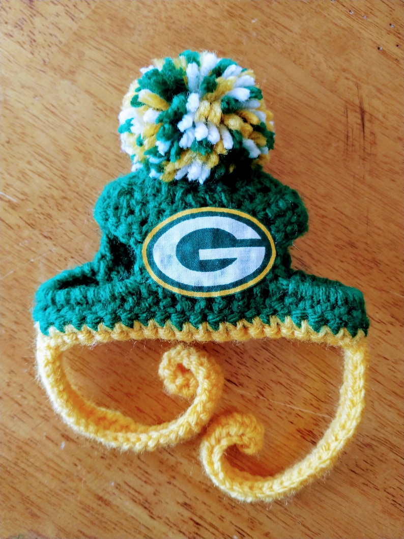 May include: A handmade, green and yellow crocheted hat with a pom-pom and the Green Bay Packers logo. The hat features a green body, yellow trim, and a white and yellow pom-pom. The logo is an oval with a white background and a green "G".
