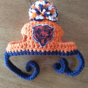Puede incluir: Un peque&ntilde;o gorro de ganchillo naranja con un borde azul oscuro y dos lazos largos y rizados de color azul oscuro. El gorro est&aacute; rematado con un pomp&oacute;n multicolor en naranja, blanco y azul oscuro, y muestra un parche del logo de los Chicago Bears.