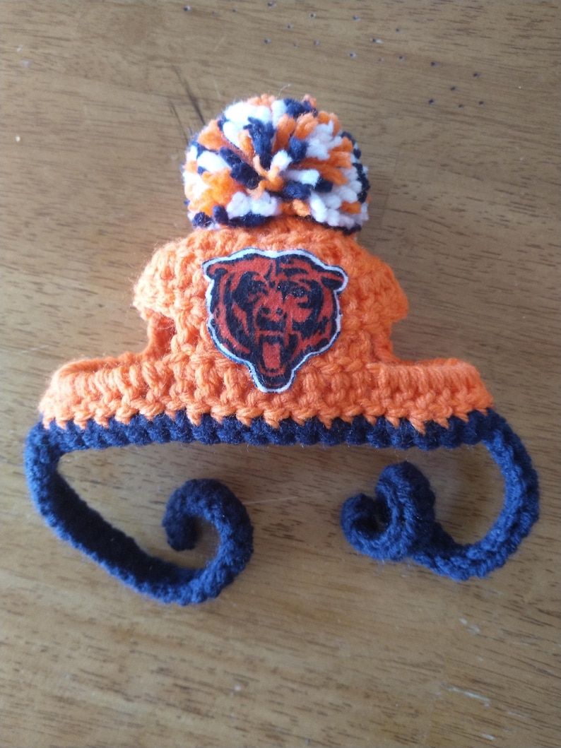 Puede incluir: Un peque&ntilde;o gorro de ganchillo naranja con un borde azul oscuro y dos lazos largos y rizados de color azul oscuro. El gorro est&aacute; rematado con un pomp&oacute;n multicolor en naranja, blanco y azul oscuro, y muestra un parche del logo de los Chicago Bears.