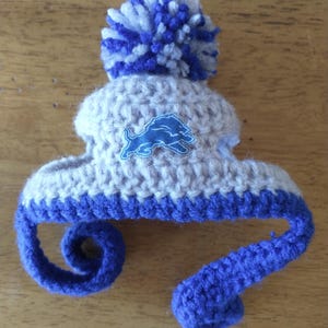 Puede incluir: Un peque&ntilde;o gorro de ganchillo en hilo color canela claro y azul real, con un gran pomp&oacute;n en la parte superior y dos largas cintas trenzadas azules colgantes. Un parche bordado azul que representa un le&oacute;n de perfil est&aacute; centrado al frente.