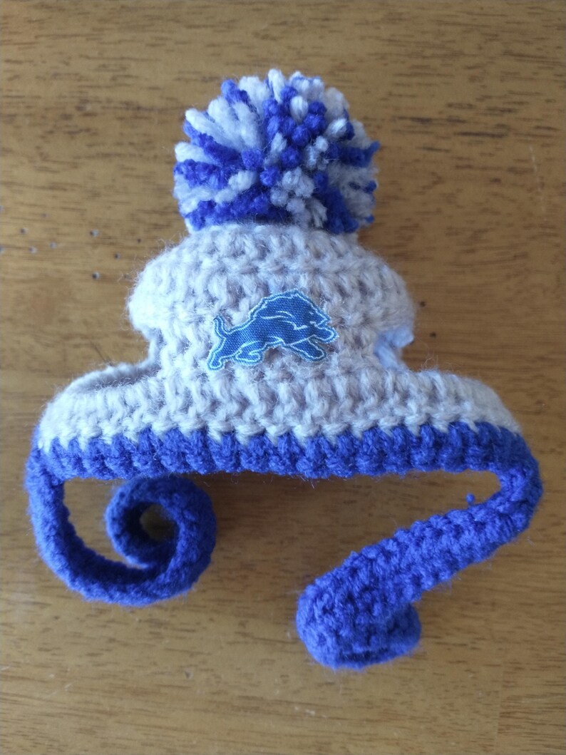 Puede incluir: Un peque&ntilde;o gorro de ganchillo en hilo color canela claro y azul real, con un gran pomp&oacute;n en la parte superior y dos largas cintas trenzadas azules colgantes. Un parche bordado azul que representa un le&oacute;n de perfil est&aacute; centrado al frente.