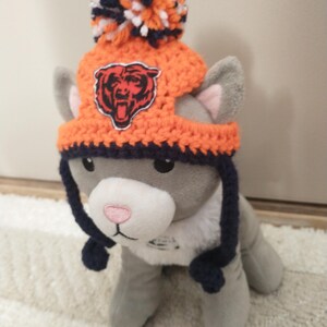 May include: A gray plush cat wearing an orange and navy blue crocheted hat with a pom-pom and a Chicago Bears logo. The hat has ear flaps and a navy blue brim. The cat has a pink nose and is sitting on a white rug.