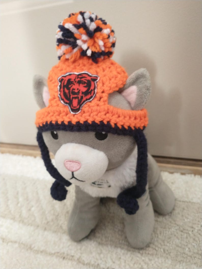 May include: A gray plush cat wearing an orange and navy blue crocheted hat with a pom-pom and a Chicago Bears logo. The hat has ear flaps and a navy blue brim. The cat has a pink nose and is sitting on a white rug.