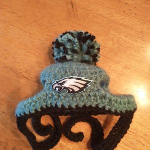 May include: Handmade crochet hat in teal and black, featuring a pom-pom and the Philadelphia Eagles logo. The hat has earflaps and is designed for warmth and style. A unique accessory for fans.