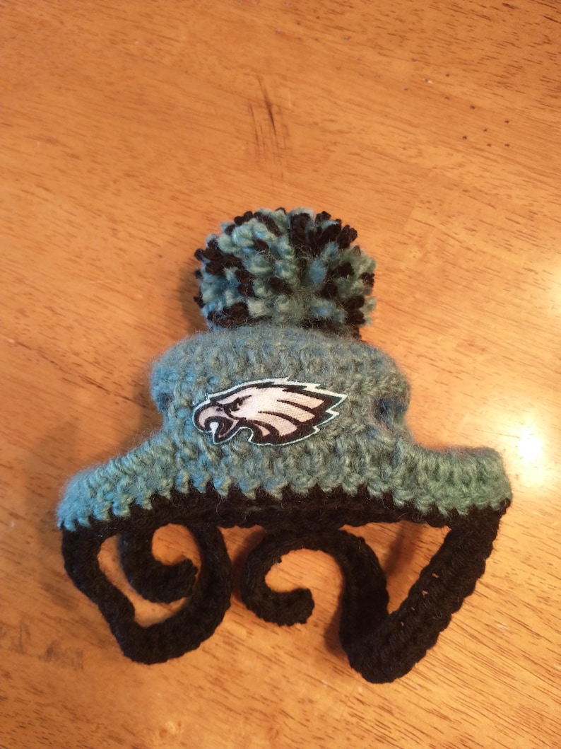 May include: Handmade crochet hat in teal and black, featuring a pom-pom and the Philadelphia Eagles logo. The hat has earflaps and is designed for warmth and style. A unique accessory for fans.