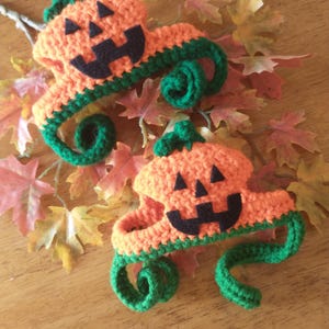 May include: Two crocheted pumpkin hats with green stems and orange bodies. The pumpkins have black felt faces with a smiling expression.