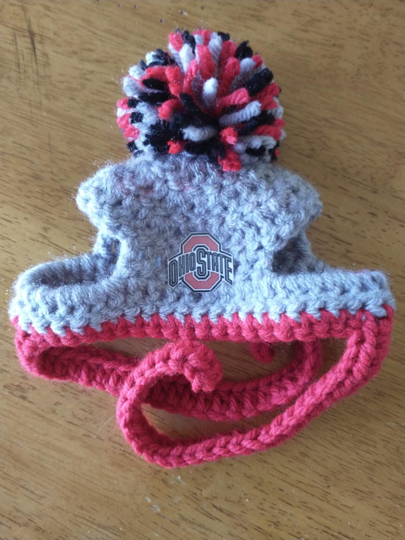 May include: A gray and red crocheted pet hat with a pom-pom on top. The hat features the Ohio State logo and a red strap. The pom-pom is made of red, black, and white yarn.