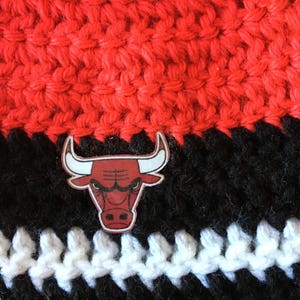 Pode incluir: Um chap&eacute;u de croch&ecirc; preto e vermelho com uma faixa branca e um logotipo de cabe&ccedil;a de touro vermelho e preto.