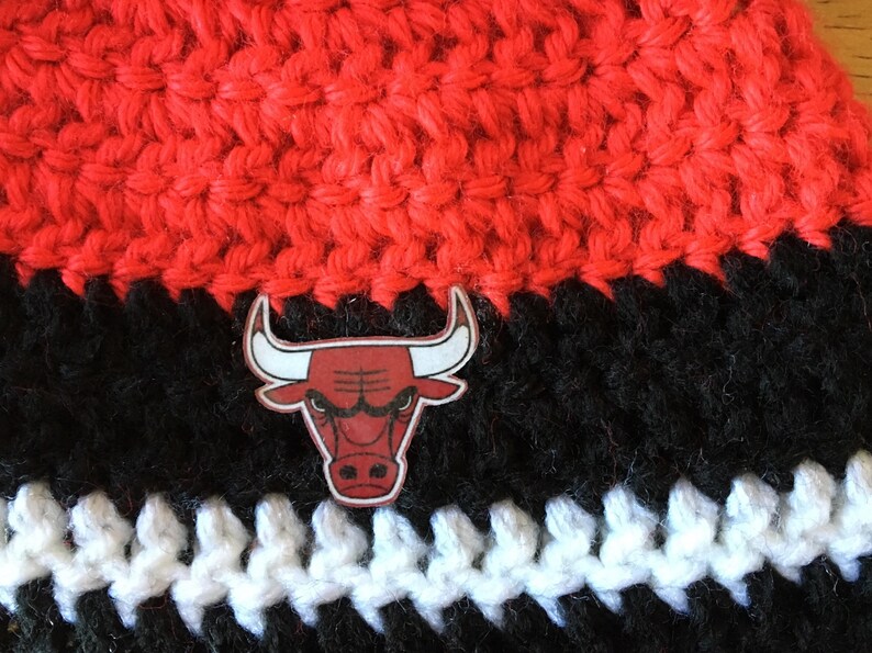 Pode incluir: Um chap&eacute;u de croch&ecirc; preto e vermelho com uma faixa branca e um logotipo de cabe&ccedil;a de touro vermelho e preto.