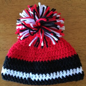 Pode incluir: Chap&eacute;u de beb&eacute; em croch&ecirc; vermelho, preto e branco com um pompom no topo.