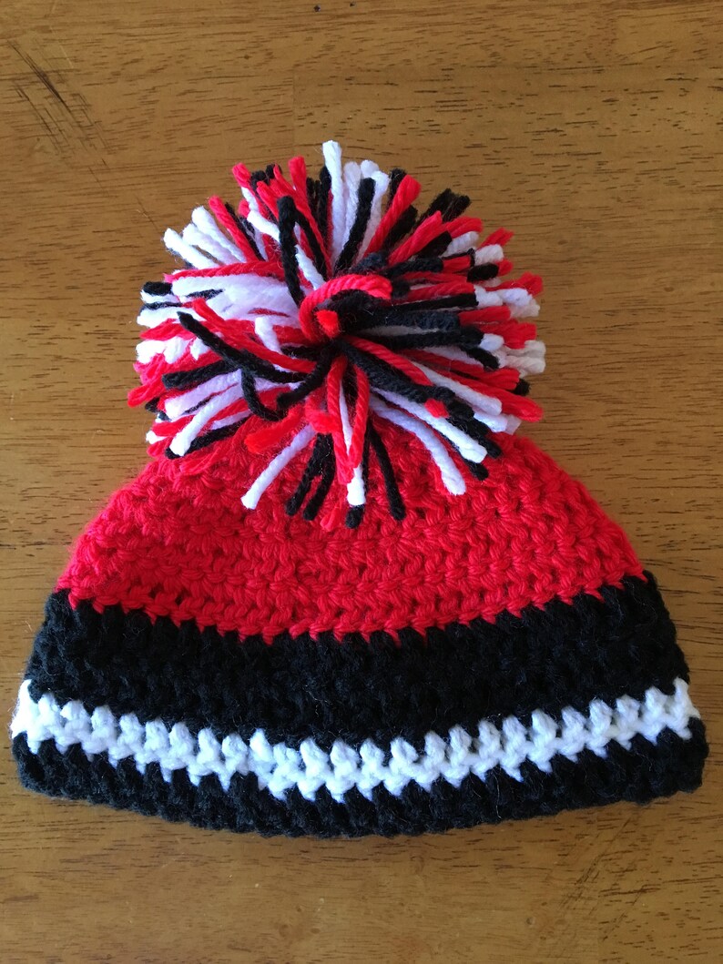 Pode incluir: Chap&eacute;u de beb&eacute; em croch&ecirc; vermelho, preto e branco com um pompom no topo.
