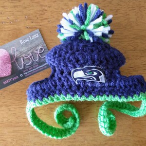 May include: A handmade, blue and green crocheted hat with a pom-pom. The hat features the Seattle Seahawks logo and ear flaps. A business card with the word "love" and a pink heart is next to the hat.