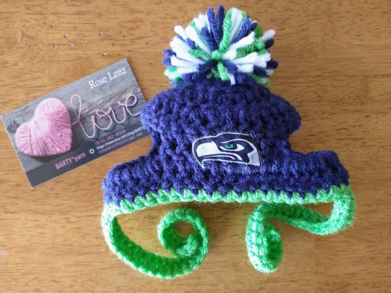May include: A handmade, blue and green crocheted hat with a pom-pom. The hat features the Seattle Seahawks logo and ear flaps. A business card with the word "love" and a pink heart is next to the hat.