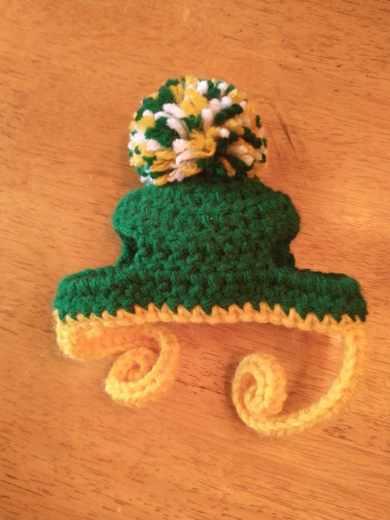 Puede incluir: Un peque&ntilde;o gorro de ganchillo en verde oscuro con un borde amarillo y dos lazos amarillos rizados que cuelgan de los lados. La parte superior del gorro est&aacute; adornada con un gran pomp&oacute;n multicolor con hebras de lana verde, amarilla y blanca.