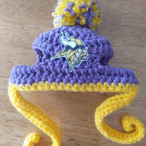 May include: A handmade purple and yellow crochet hat with a pom-pom and ear flaps. The hat features the Minnesota Vikings logo on the front. The hat is made of yarn and is designed for a baby or small child.