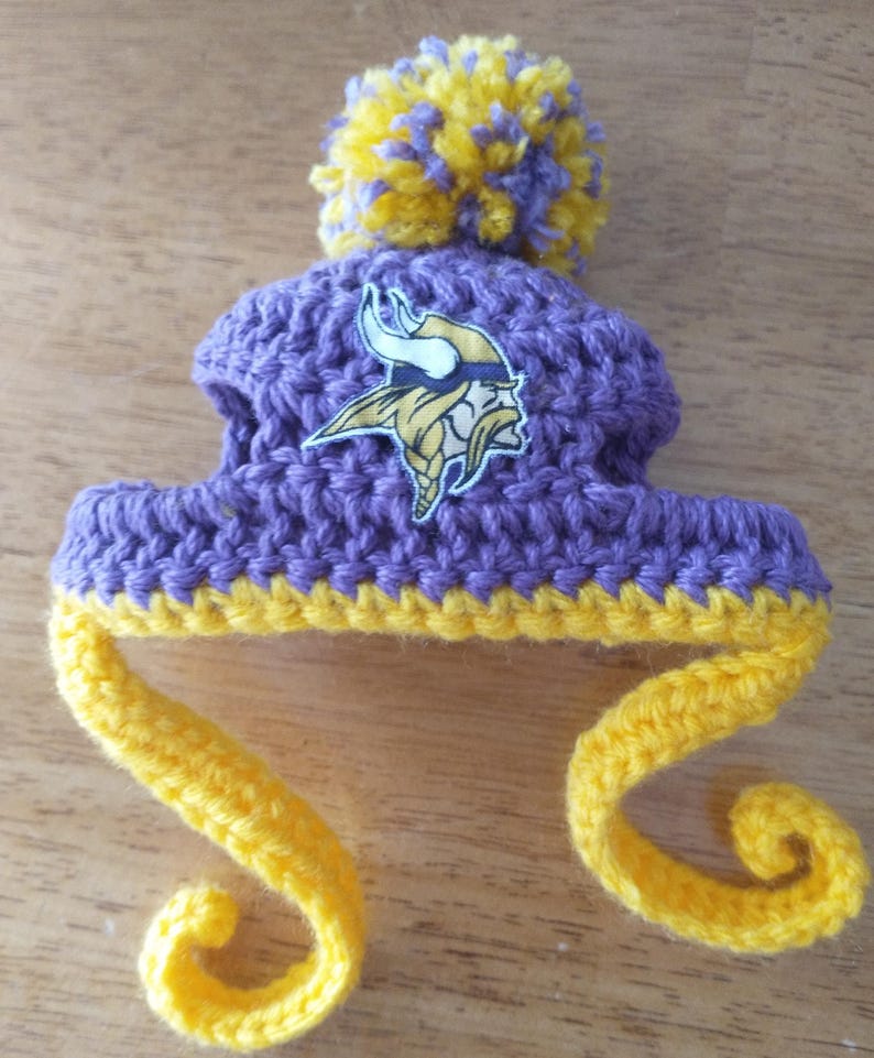 May include: A handmade purple and yellow crochet hat with a pom-pom and ear flaps. The hat features the Minnesota Vikings logo on the front. The hat is made of yarn and is designed for a baby or small child.