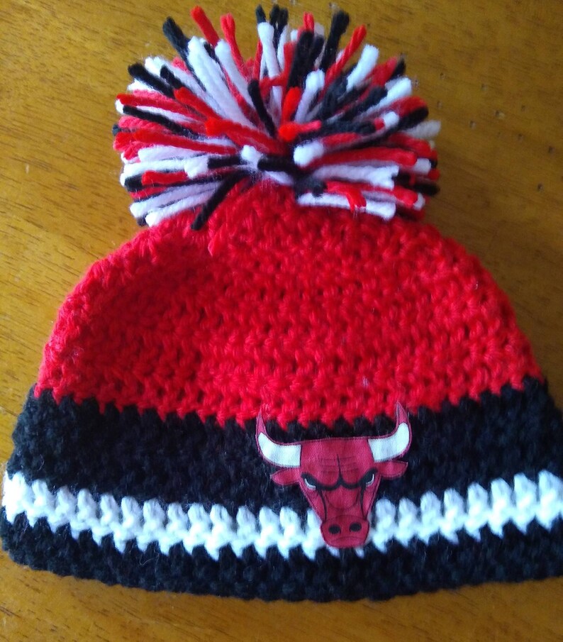 Pode incluir: Boina de croch&ecirc; vermelha e preta com um pompom branco e um logotipo de touro vermelho e preto.