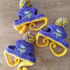 May include: Three handmade crochet hats in purple and yellow, featuring a pom-pom on top and a curled brim. Each hat has a logo of a Viking head. The hats are displayed on a wooden surface.
