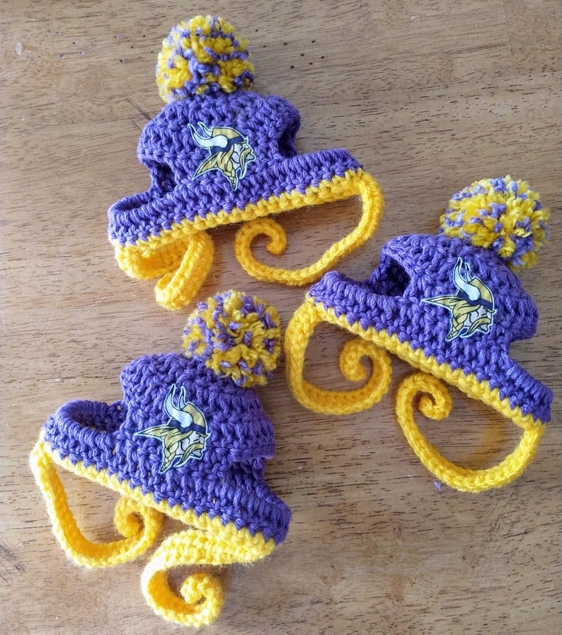 May include: Three handmade crochet hats in purple and yellow, featuring a pom-pom on top and a curled brim. Each hat has a logo of a Viking head. The hats are displayed on a wooden surface.