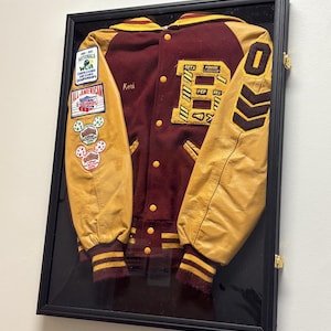 May include: A framed maroon and gold varsity jacket. The jacket has gold leather sleeves, a maroon body, and patches. The letter "B" and the number "0" are visible. The name "Keri" is embroidered on the jacket. The jacket is displayed in a black frame.