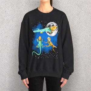 Op de afbeelding: Zwarte sweatshirt met een fantasie-illustratie van drie hagedissen in een nachtelijke hemel. Eén hagedis zweeft in de buurt van een volle maan, de andere eronder. De hagedissen zijn in tinten blauw, geel en oranje, tegen een blauwe en sterrenhemel.