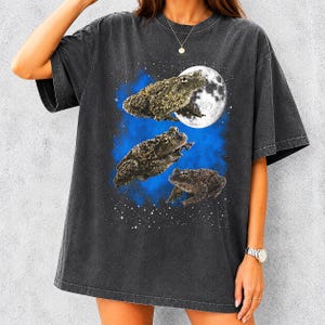 Può includere: T-shirt grigio scuro con un design stravagante di tre rospi su uno sfondo blu e stellato, con una luna piena. I rospi sono dettagliati con la pelle strutturata, creando una maglietta grafica unica e accattivante.