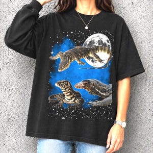Puede incluir: Camiseta negra con un diseño gráfico de tres varanos sobre un fondo azul estrellado y una luna llena. Los varanos están representados con escamas y marcas detalladas en varias poses.