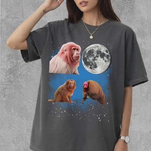 May include: Dark gray t-shirt featuring a graphic design of three monkeys, a full moon, and a starry sky. The monkeys are depicted in shades of pink and brown against a blue background. The shirt is a casual, oversized fit.