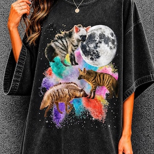 Può includere: T-shirt nero con un design grafico di tre iene, una luna piena e schizzi di vernice colorati. Le iene sono raffigurate in varie pose, una sembra annusare la luna. Il design è ambientato su uno sfondo stellato.