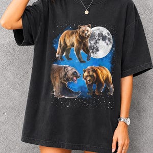 Könnte beinhalten: Ein schwarzes Kurzarm-T-Shirt mit einem Grafikdruck von drei Braunbären vor einem blauen Sternenhimmel mit einem großen Vollmond. Die Bären sind in verschiedenen Posen dargestellt, darunter stehend, knurrend und gehend.